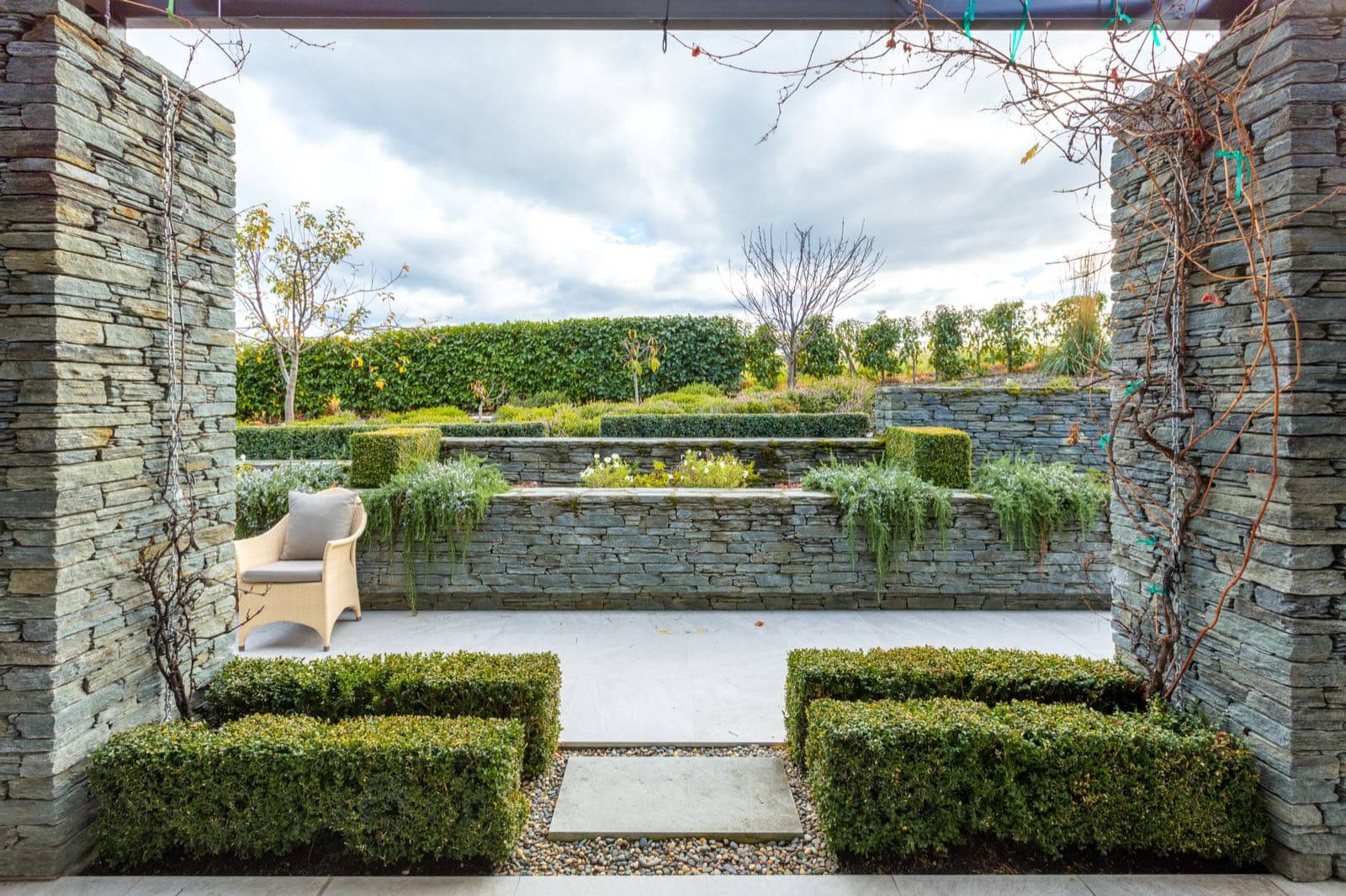 A walled schist courtyard with clipped box hedging, a seat and an orchard beyond