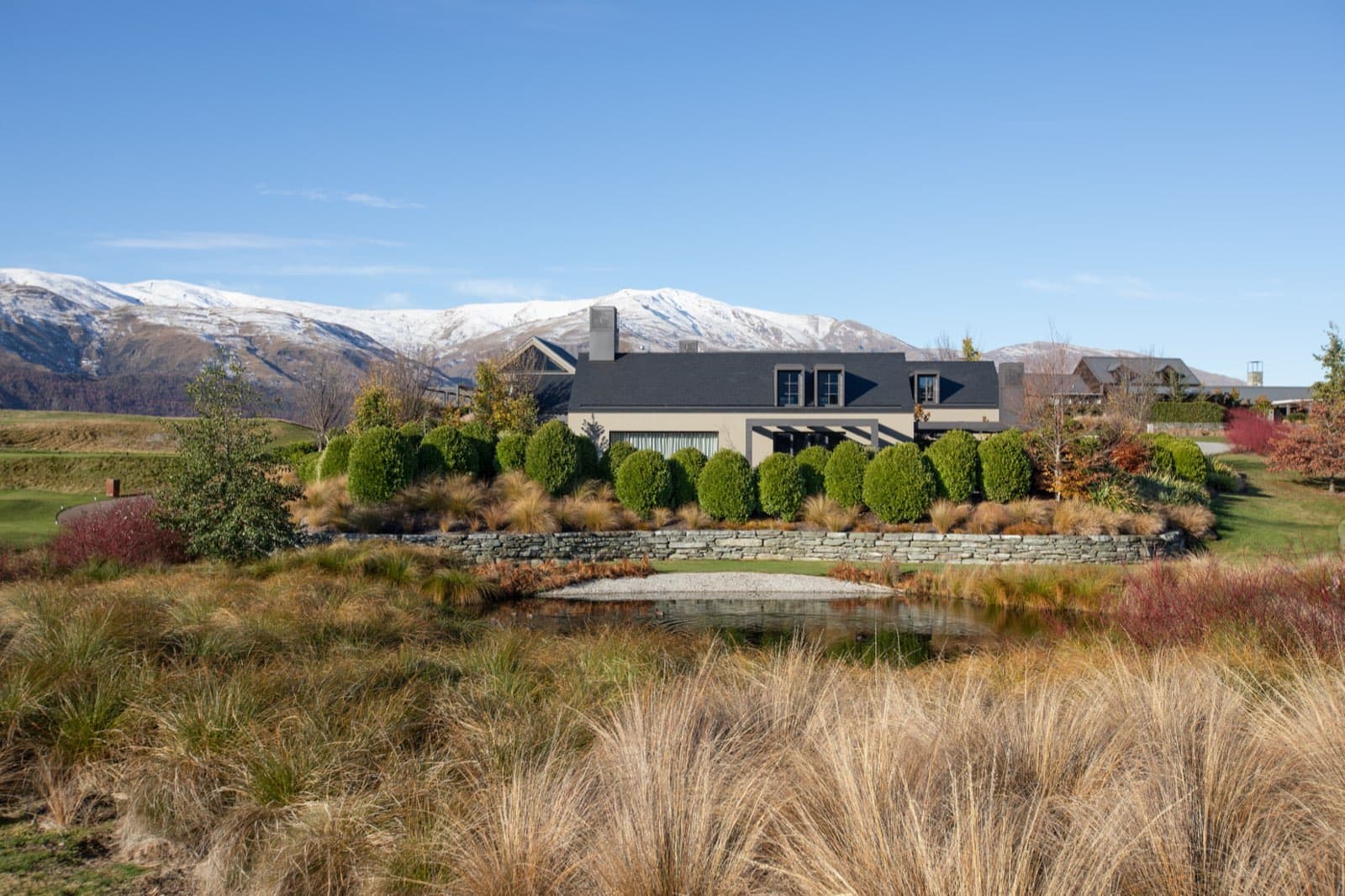 Millbrook Sanctuary across its pond, with tussock planting, a schist wall and snow on the range behind