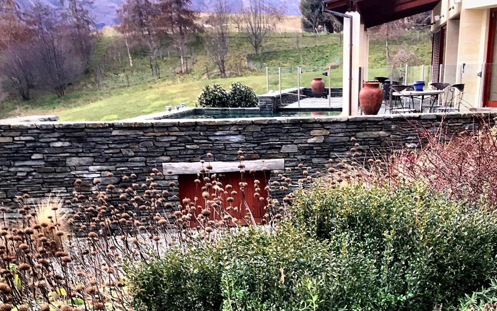 A schist-walled terrace garden overgrown, with loose hedges and spent perennials before renovation