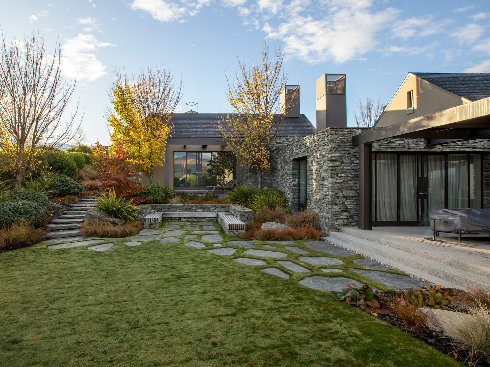 A stone house above a flagstone path set into lawn, autumn trees turning in low sun behind it