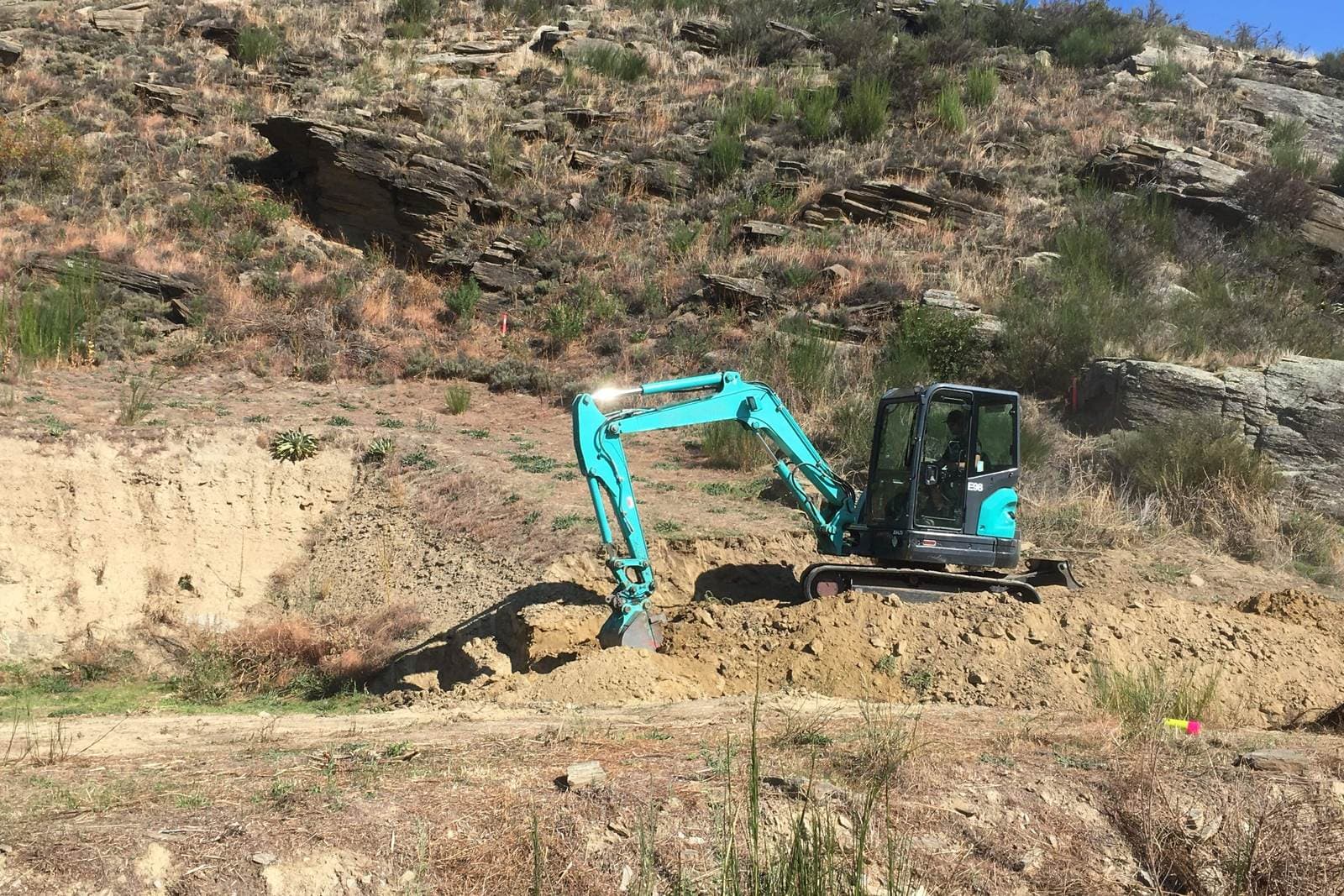 A small excavator cutting into a dry schist bank to shape the ground
